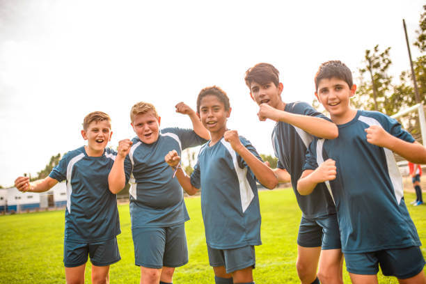 Five blue jersey preteen and teenage boys looking at camera while cheering victory on sports field.