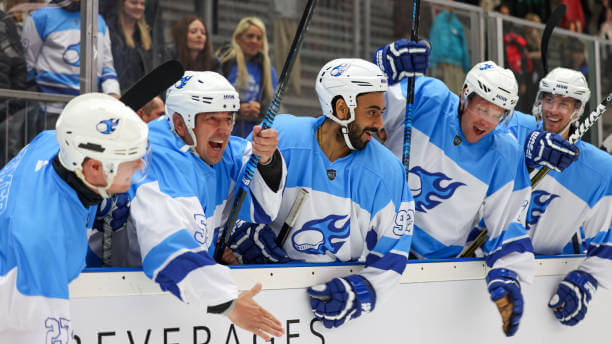 A group of joyful hockey players wearing matching blue and white uniforms celebrate enthusiastically on the bench, their energy palpable. They are congratulating each other during a pivotal moment in the game, fully engaged and exhilarated by their teamwork.